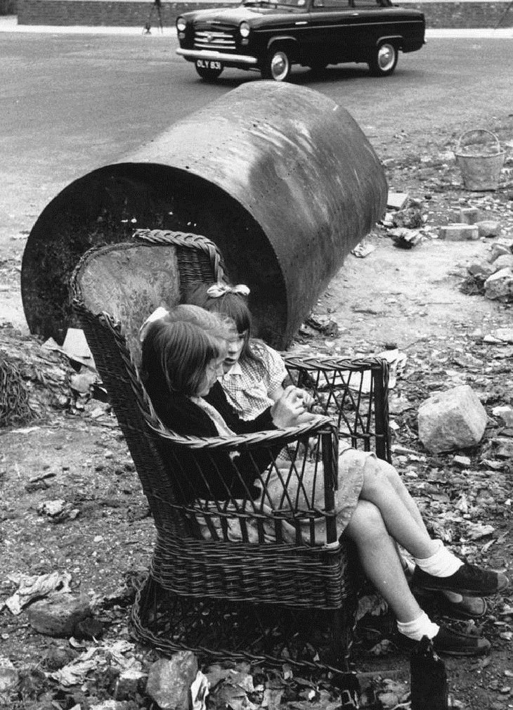 Niños Jugando En Las Calles De Londres Dos niñas sentadas en una vieja silla de mimbre junto a la carretera
