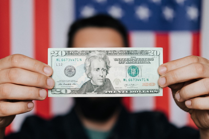 Los mejores países para el hombre jubilado que sostiene un billete de 20 dólares frente a la bandera de Estados Unidos