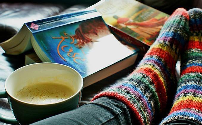 Las piernas de una mujer con calcetines de lana y un libro y una bebida caliente al lado.