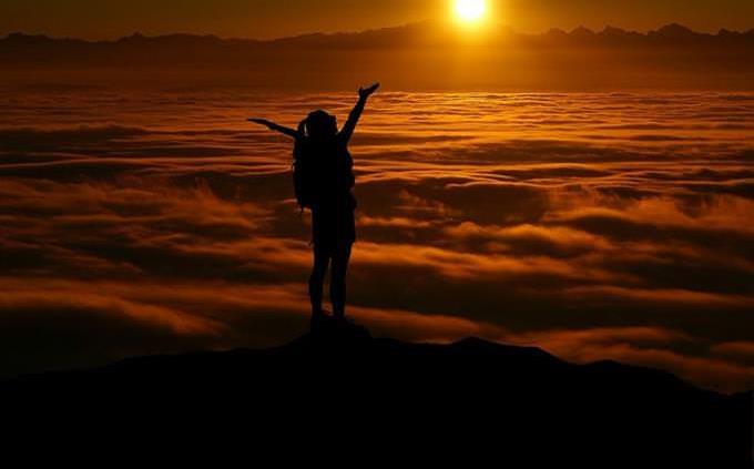 Una mujer parada sobre un acantilado y agitando sus manos en el aire durante la puesta de sol